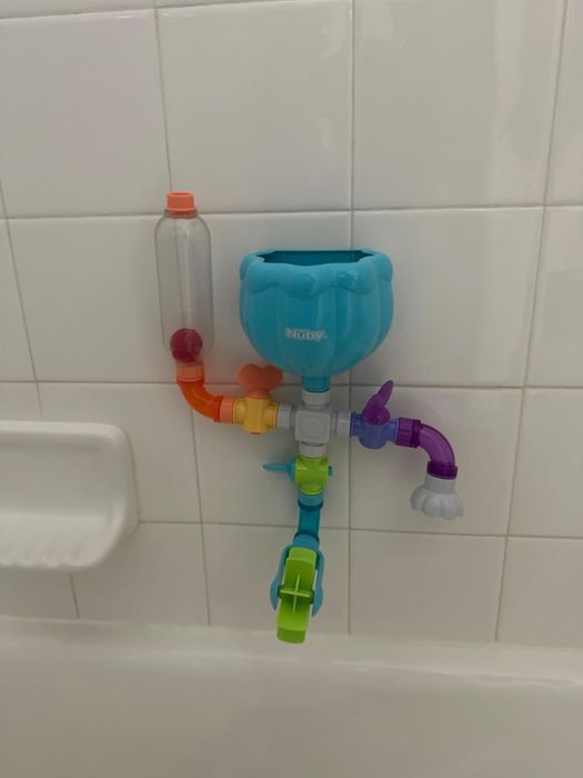 A Nuby water spout bath toy mounted on white bathroom tiles, with colorful channels and interactive features for playtime fun