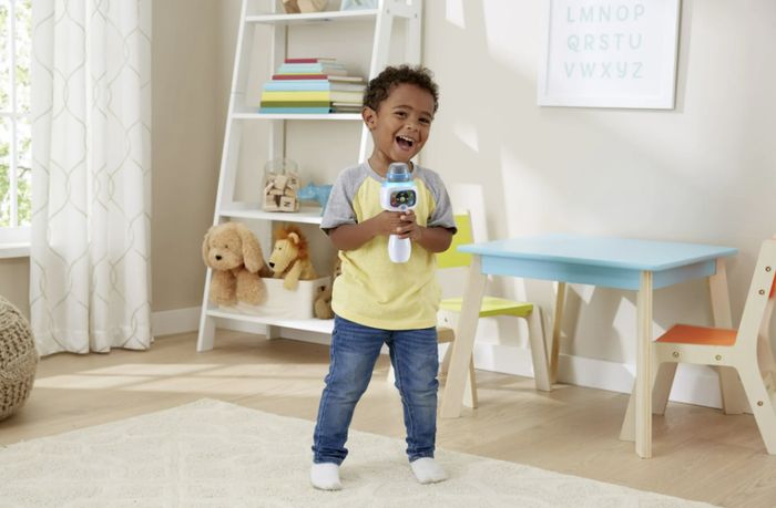 A toddler stands with joy, holding a toy microphone in a colorful playroom with a table, chairs, stuffed animals, and a wall alphabet chart.