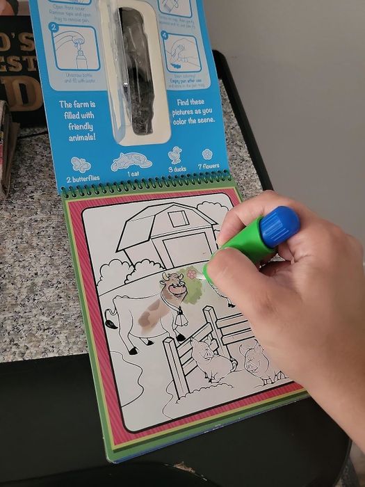 A hand using a green marker to color in barn animals on a farm-themed children's coloring book.