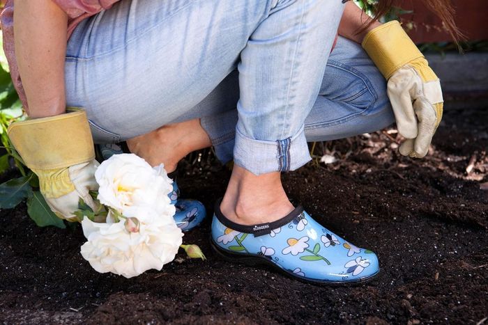 A model shows off blue floral gardening clogs, paired with jeans and gloves, while holding a white flower and squatting in the soil.