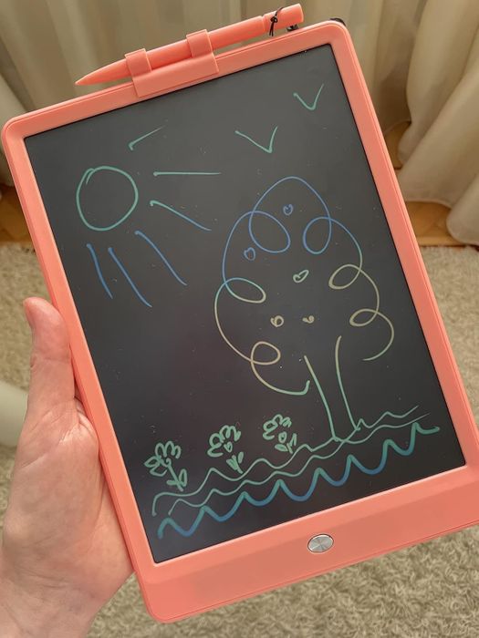 A hand holds an LCD writing tablet displaying a colorful drawing of a sun, tree, birds, and flowers by a child