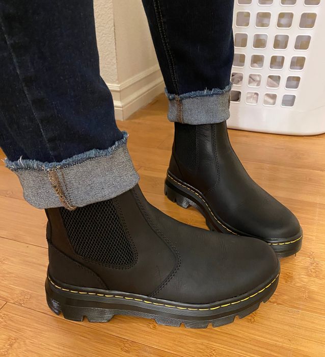 A reviewer is seen wearing black ankle boots with yellow stitching, paired with dark jeans rolled up at the ankles for a casual, stylish look.