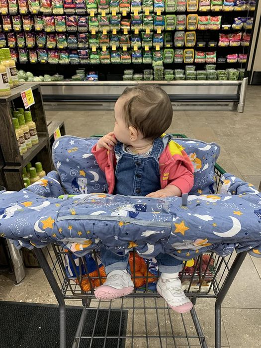 a baby in a shopping cart, wearing a pink jacket and denim overalls, looking to the side in a grocery store’s produce aisle