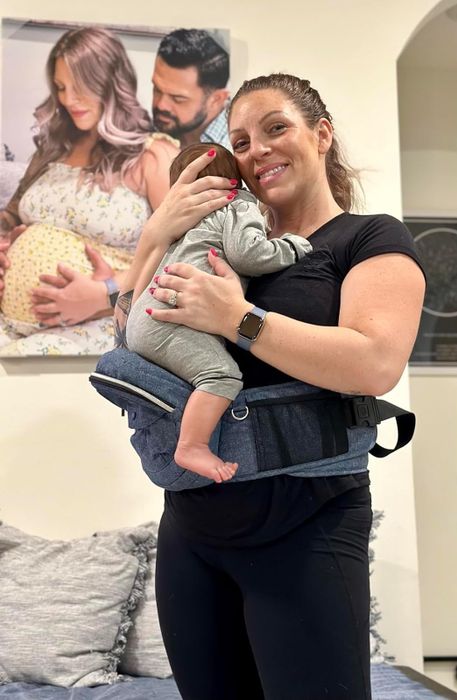 a smiling individual uses a hip seat carrier to comfortably hold their baby
