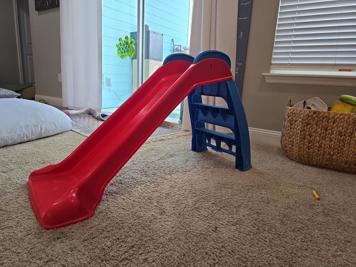 A small red plastic slide with a blue ladder, placed on a carpeted floor in a playroom near a window. A basket and some toys are visible in the background