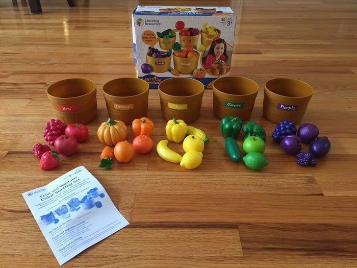 A toy set featuring five brown bowls marked with different colors, a variety of plastic fruits and veggies, along with a pamphlet and packaging showcasing a young girl playing with the set