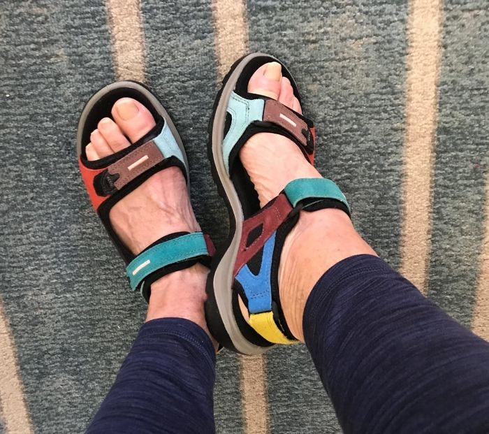 A reviewer shows off sporty, multi-colored Ecco Yucatan sandals with blue leggings, standing on a striped carpet for a fun, active vibe.