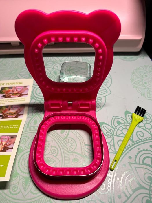 a waffle maker shaped like a bear face, featuring a double grill and a cleaning brush, with the user manual visible nearby