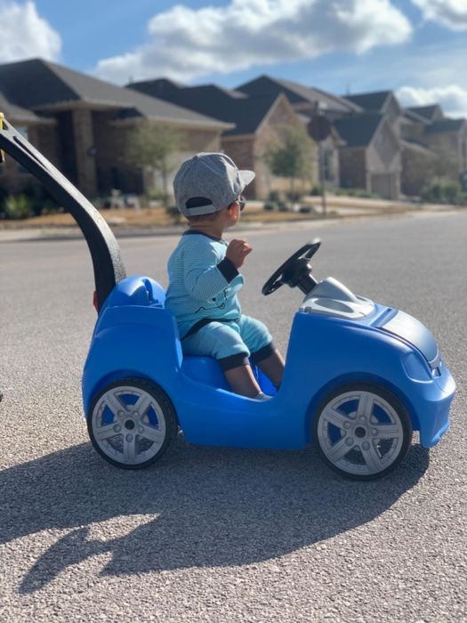 A young child in a cap and light clothes sits in a toy car, being pushed along a suburban street by an unseen adult.
