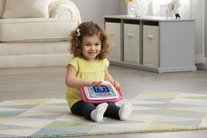 A child smiles while seated on a rug, enjoying a toy tablet in the living room.