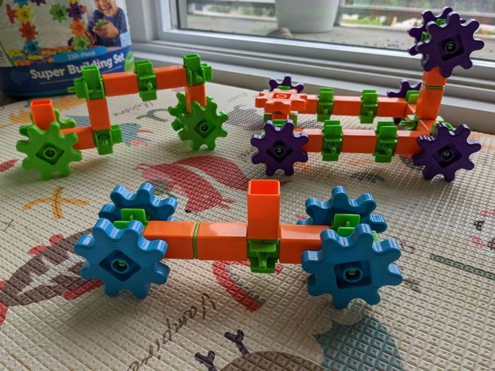 A variety of colorful toy vehicles made of interlocking blocks with gears, set on a play mat near a window. A box labeled 'Super Building Set' sits in the background.