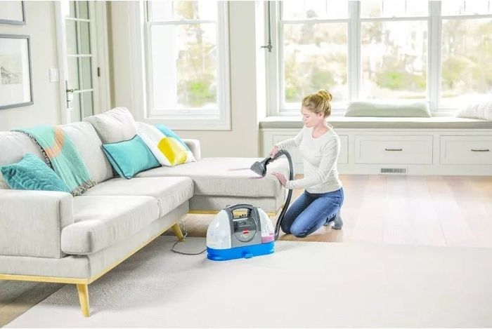 A person kneeling in a living room uses a small carpet cleaner on a piece of furniture. The room includes a sofa, colorful pillows, and large windows.