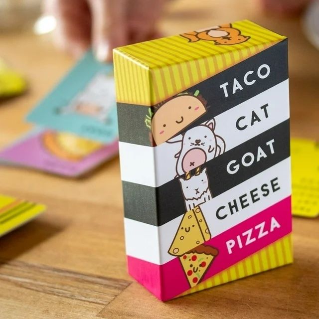 The 'Taco Cat Goat Cheese Pizza' card game box is shown on a wooden table, with colorful cards spread out around it. Hands can be seen in the background as players prepare for their next round.
