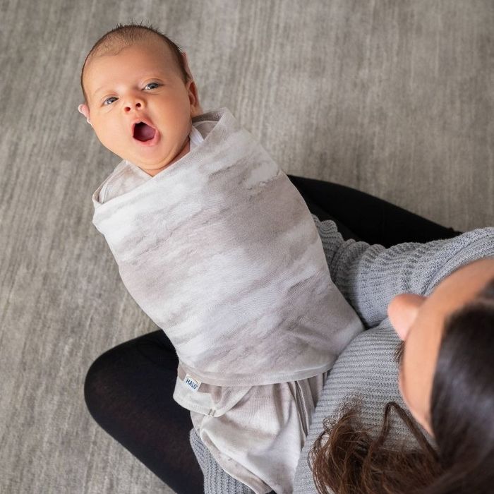 A baby wrapped in a swaddle blanket, yawning, while being held by an adult in a sweater, viewed from above