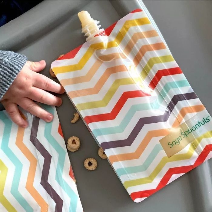 A child’s hand holding a multicolored chevron-patterned food pouch with cereal scattered around. The pouch reads: Sage Spoonfuls