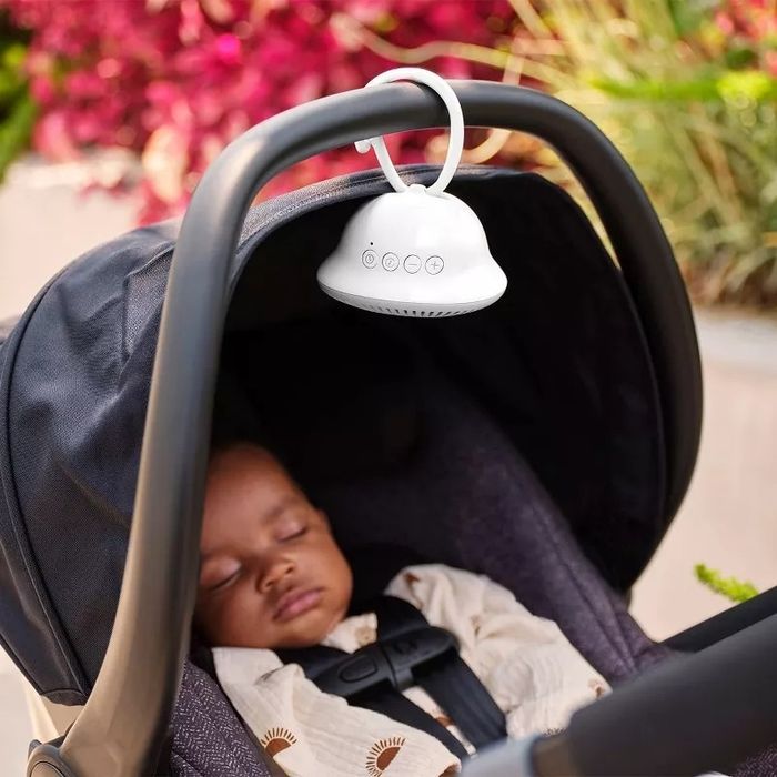 A baby sleeping in a stroller with a white noise machine clipped to the handle