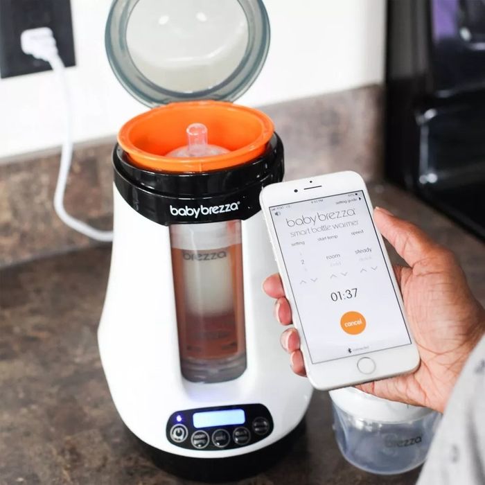 A person using a smartphone to control a Baby Brezza Formula Pro machine placed on a kitchen counter
