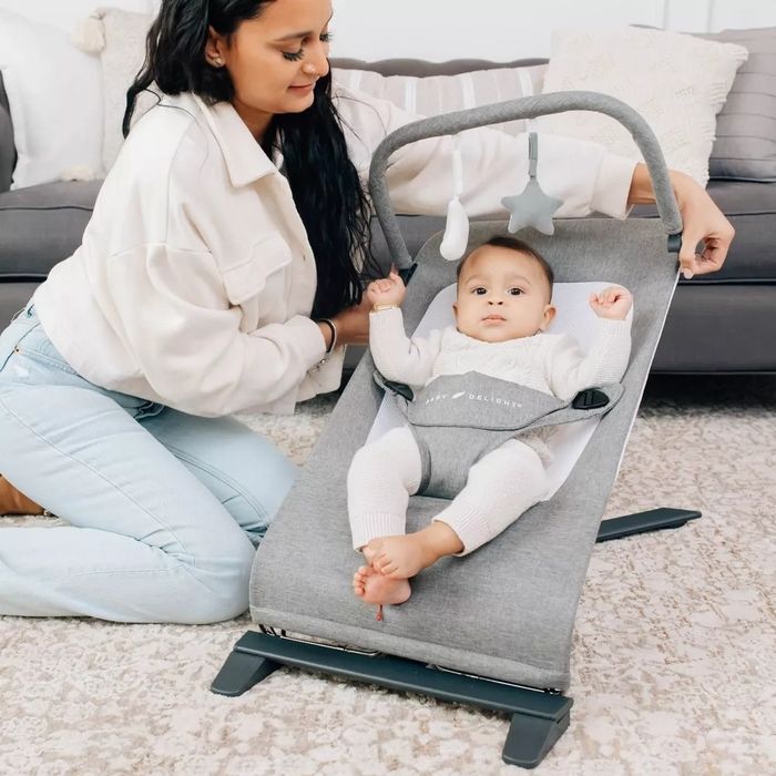A model kneeling next to a baby sitting in a gray bouncer with star and cloud-shaped toys