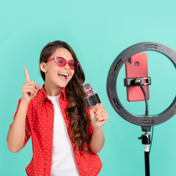 A young model with long hair and glasses, dressed casually, holding a microphone and pointing upwards, in front of a ring light with a smartphone mounted.