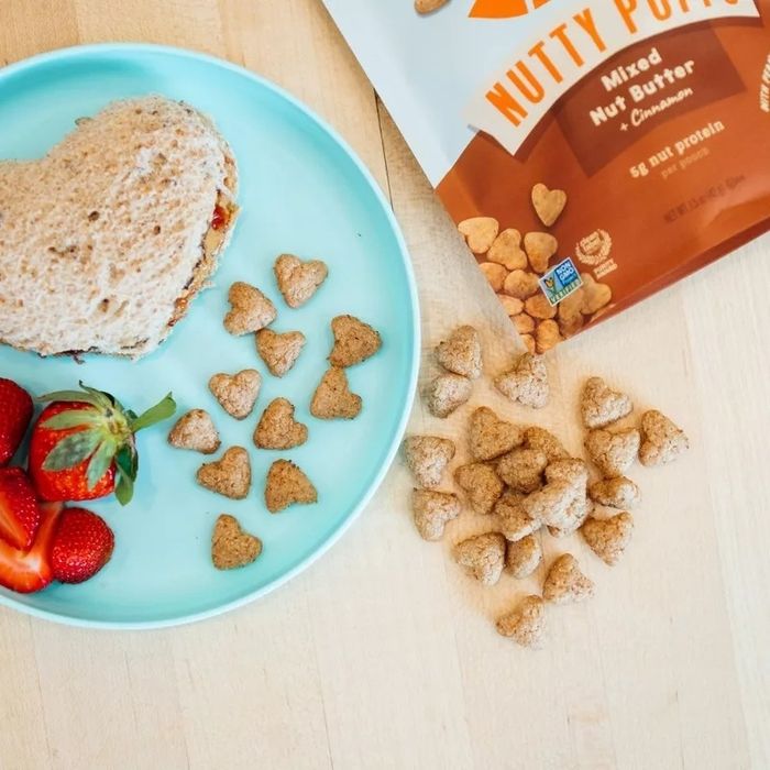 Heart-shaped nut butter snacks arranged with a heart-shaped sandwich and strawberries on a plate. A Nutty Pops package is visible, set in a shopping-themed scene.