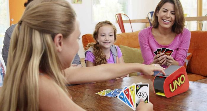 A group of three people is enjoying a lively game of Uno, complete with a card launcher. The player on the left is placing a card into the launcher, all while smiling and laughing together around a wooden table.