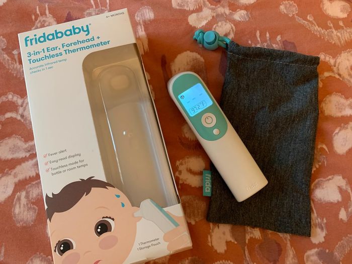 A Fridababy 3-in-1 thermometer for ear, forehead, and touchless use, complete with packaging, a storage pouch, and a visible temperature display