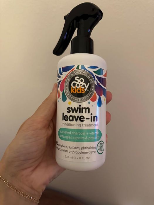 A hand holds a bottle of SoCozy Kids swim leave-in conditioner treatment, featuring activated charcoal and vitamin D, with a spray nozzle for easy and convenient application