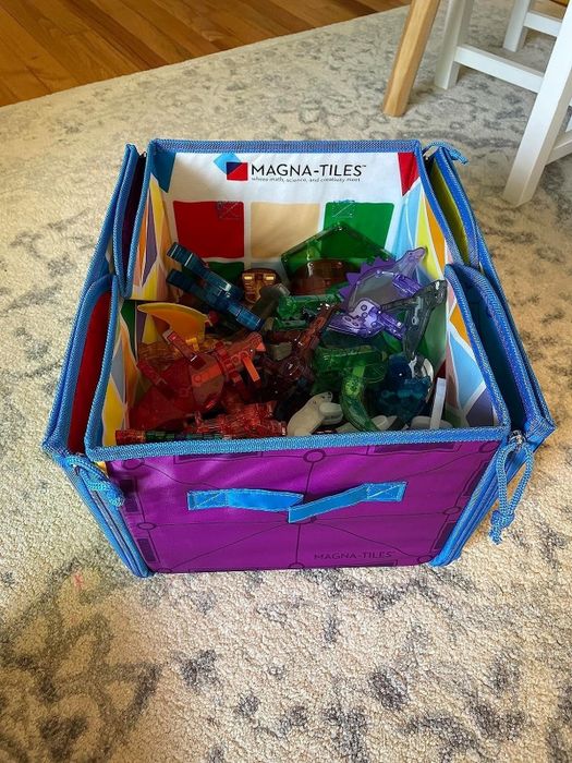 An open storage bin filled with Magna-Tiles and an assortment of colorful plastic toys scattered across a carpet.