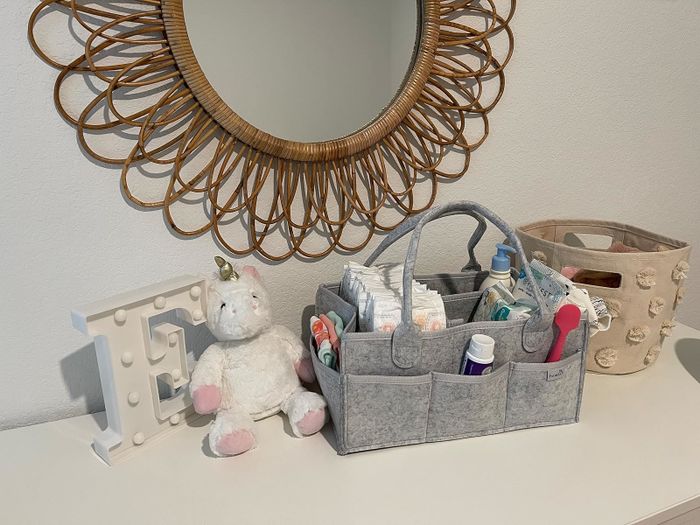 A nursery dresser with a charming decorative mirror, a plush unicorn toy, an initial letter 'E' decor, and an organized diaper caddy containing all the baby necessities.