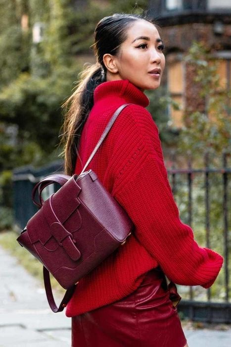 A model is showcasing a burgundy square-shaped purse with a handle on top and straps designed to be worn like a backpack.
