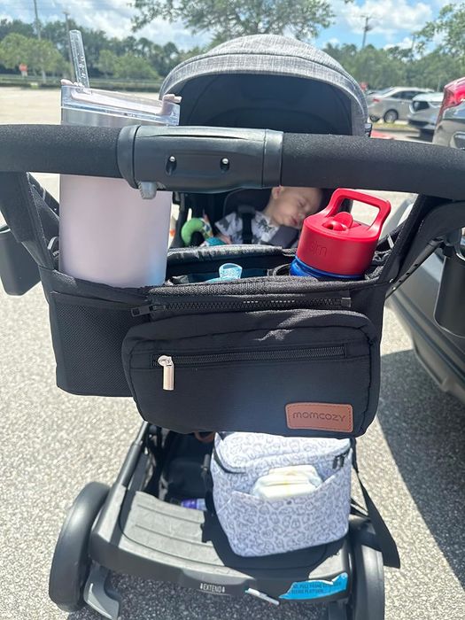 A baby sleeping peacefully in a stroller, with an organizer holding a bottle and a drink. A bag and a burp cloth are neatly tucked underneath