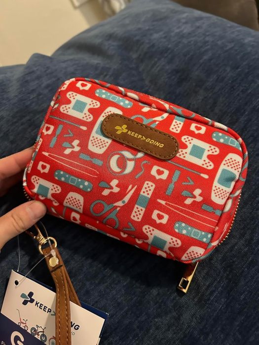 A hand holding a compact, rectangular pouch with a medical design, containing first aid essentials like bandages and scissors. The pouch is labeled