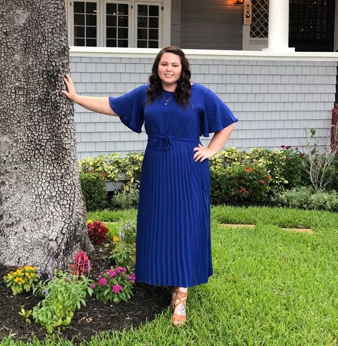 reviewer wearing the blue dress leaning against a tree