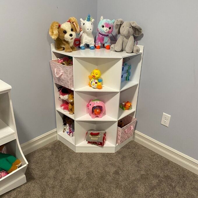 A white corner shelf, filled with a variety of children's toys including plush animals and toy vehicles, set within a room.