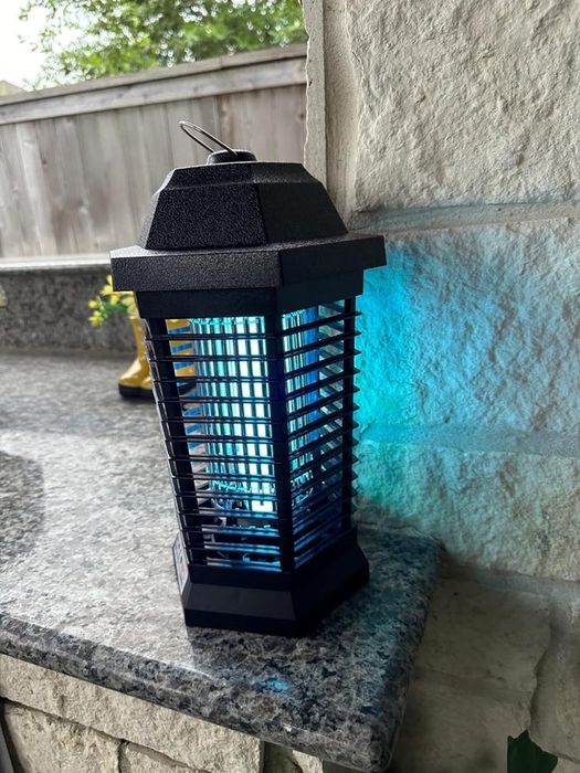 A black electric bug zapper sits outdoors on a stone countertop, glowing with a blue light.