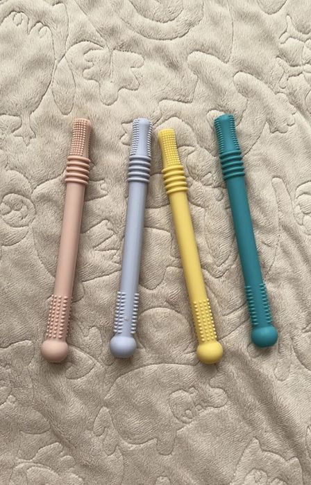 Four textured sensory chew sticks in beige, light gray, yellow, and teal, arranged side by side on a cushioned, patterned surface