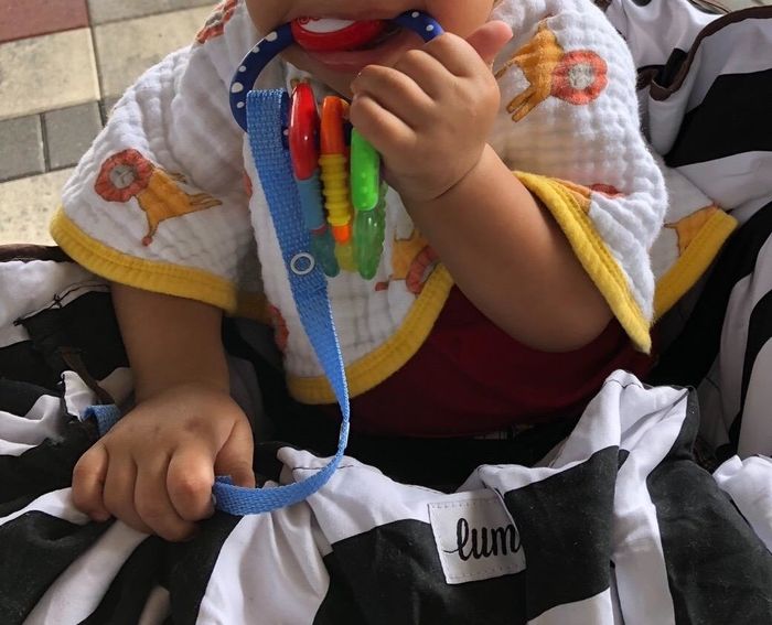 A baby in a lion-print bib, chewing on a vibrant teething toy secured by a toy strap