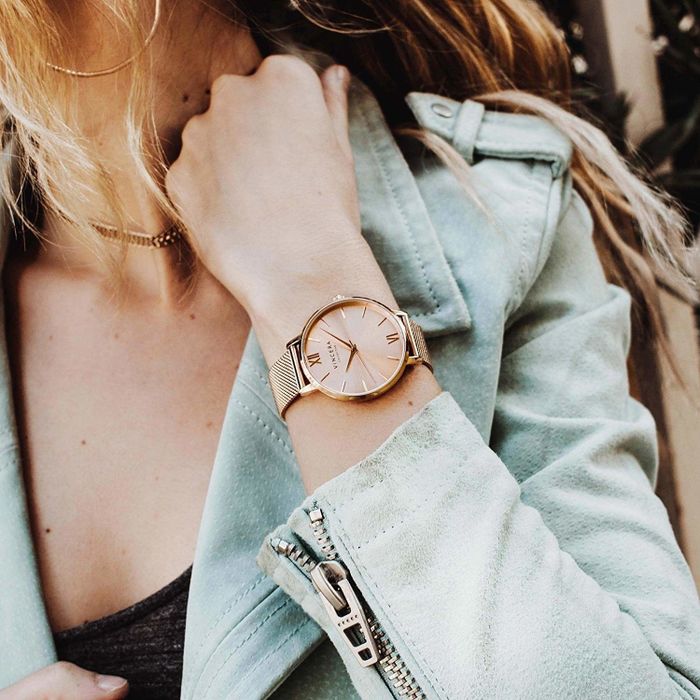 The model is wearing a round gold wristwatch with a matching gold band on their wrist.