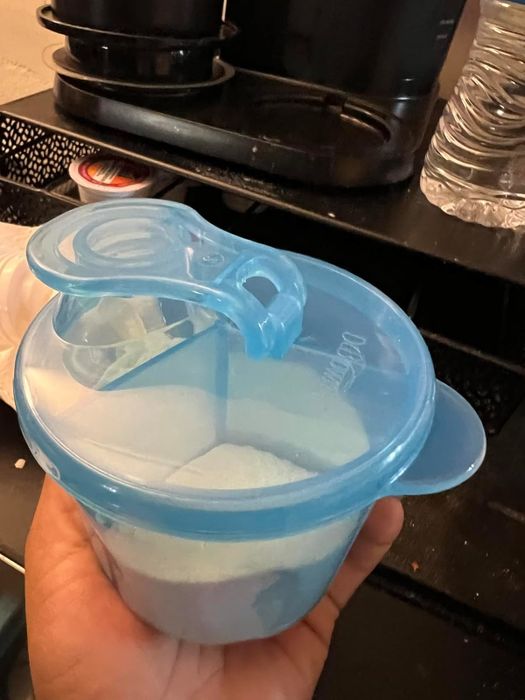 A plastic container held in hand, filled with a white powdered substance, with a coffee machine and water bottle visible in the background