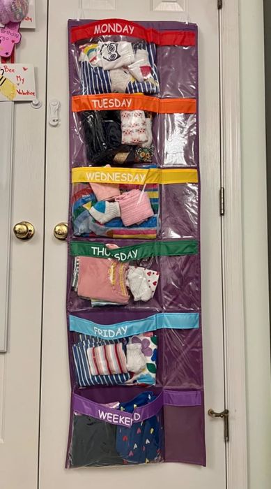 A hanging organizer with labeled compartments for each day of the week, each filled with neatly folded children's clothes and socks.