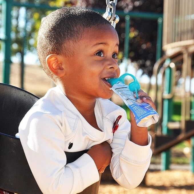 A young child happily eating from a squeezable pouch while swinging at a playground