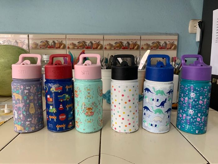 Six vibrant water bottles featuring various designs such as animals, dinosaurs, polka dots, and abstract patterns, arranged neatly on a kitchen counter