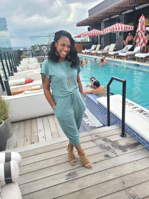 A woman in a teal casual jumpsuit and strappy sandals smiling by a poolside