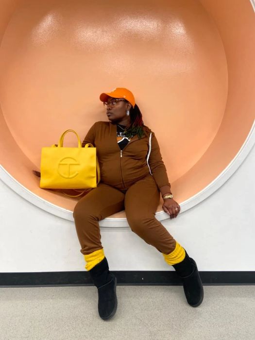 A person posing with a bright yellow tote bag, a brown outfit, and black boots against a circular backdrop