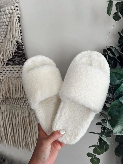 A person holding fluffy white slippers, ideal for home use, near a woven wall decoration and green plants