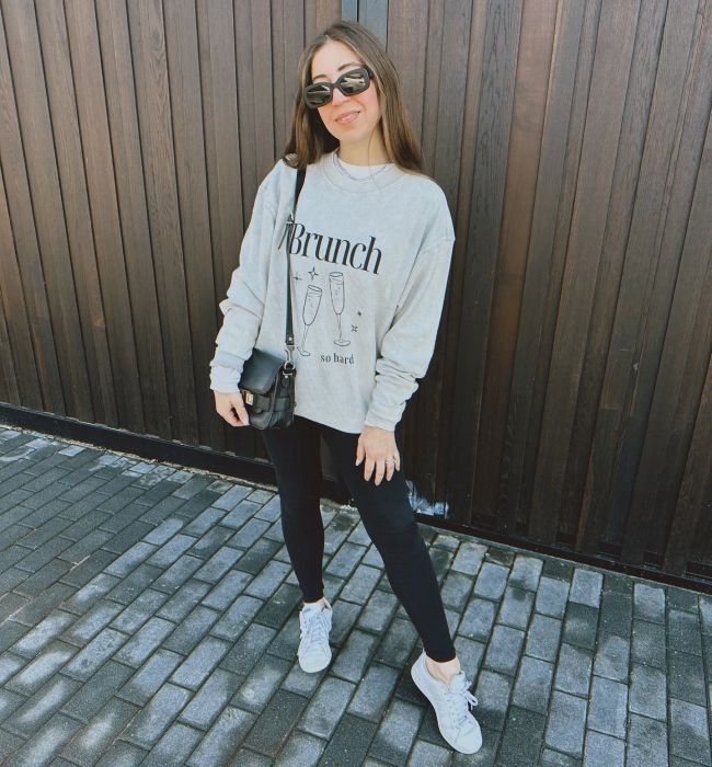 A woman smiling in a casual outfit featuring a graphic sweatshirt, black pants, sneakers, and a shoulder bag