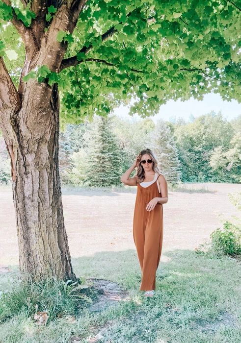 A woman in a sleeveless jumpsuit posing beside a tree, ideal for a fashion shopping feature