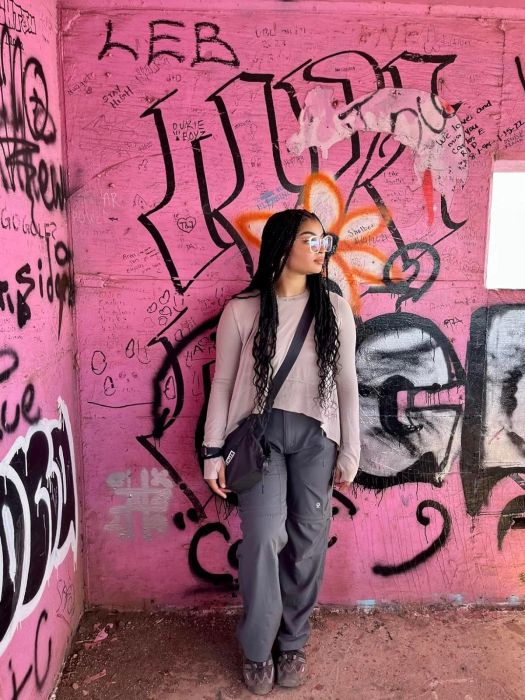 A woman with braided hair and glasses, dressed in a light top and gray pants, standing in front of a graffiti wall