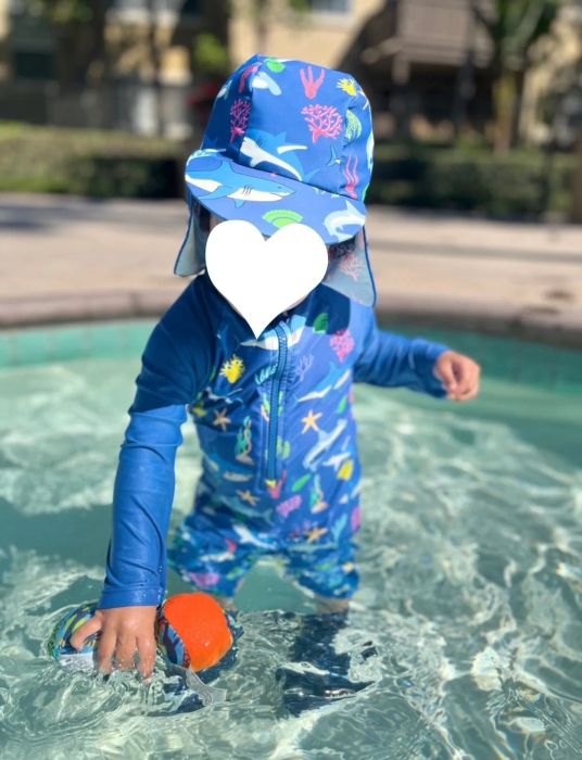 Reviewer's photo of a toddler wearing a blue aquatic-themed zip-up swimsuit, playing with a toy in shallow water. The child's face is covered with a white heart icon