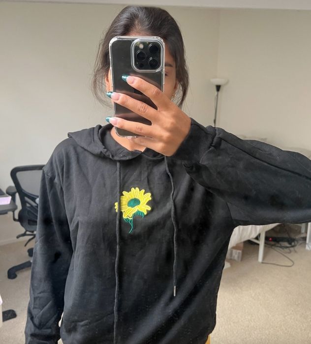 The reviewer is wearing a black hoodie with a yellow and green sunflower embroidered in the center.